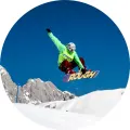 Snowboardista v zelené bundě letí nad zasněženým svahem a skalami na modré obloze, jedlý obrázek na dort