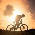 Silueta cyklisty na horském kole sjíždějícího svah proti západu slunce v oranžových a žlutých tónech, jedlý obrázek na dort